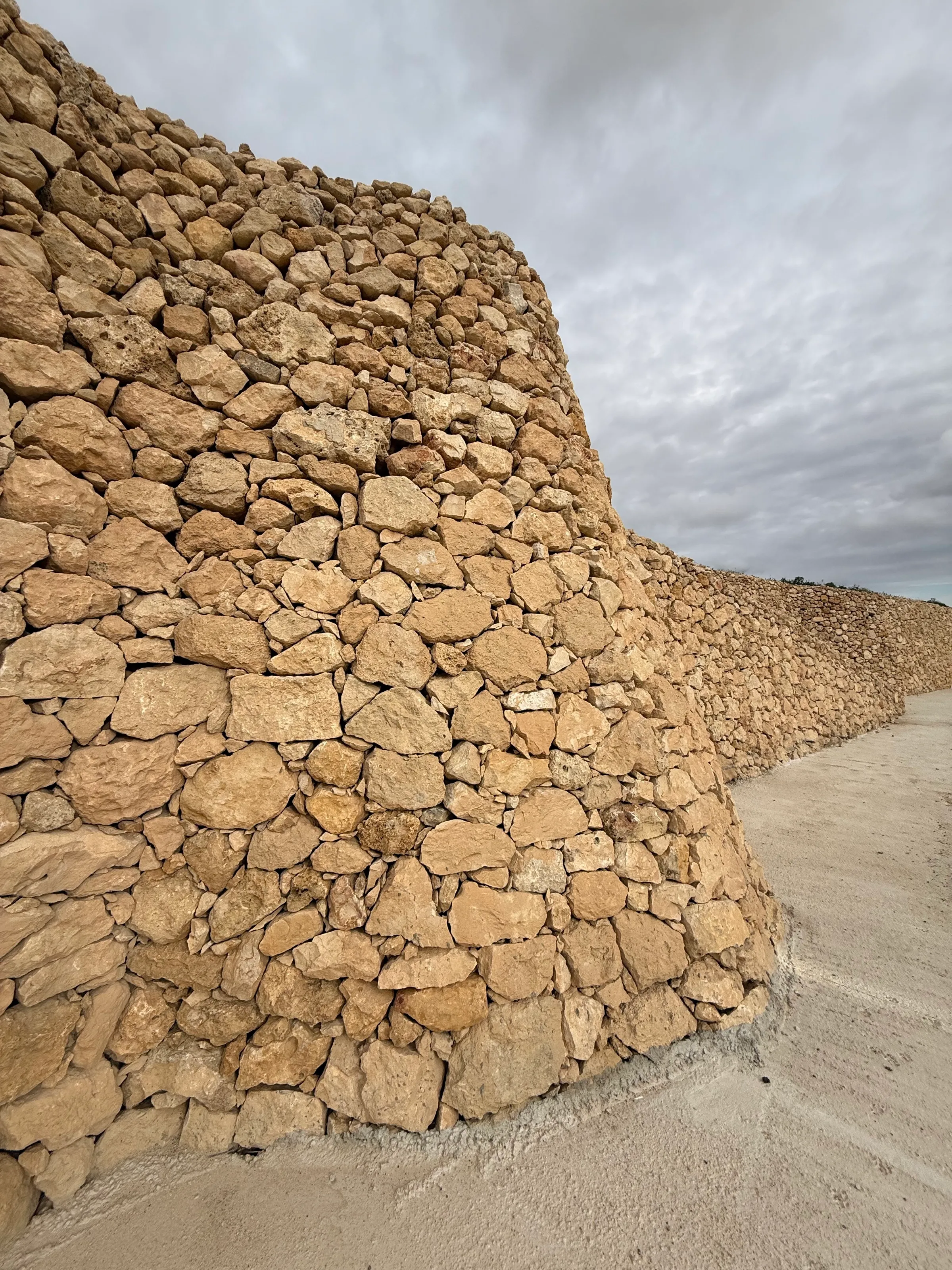 Hohe Trockenmauer aus Kalkstein, die einem Weg entlang in die Ferne führt, darüber grauer Himmel